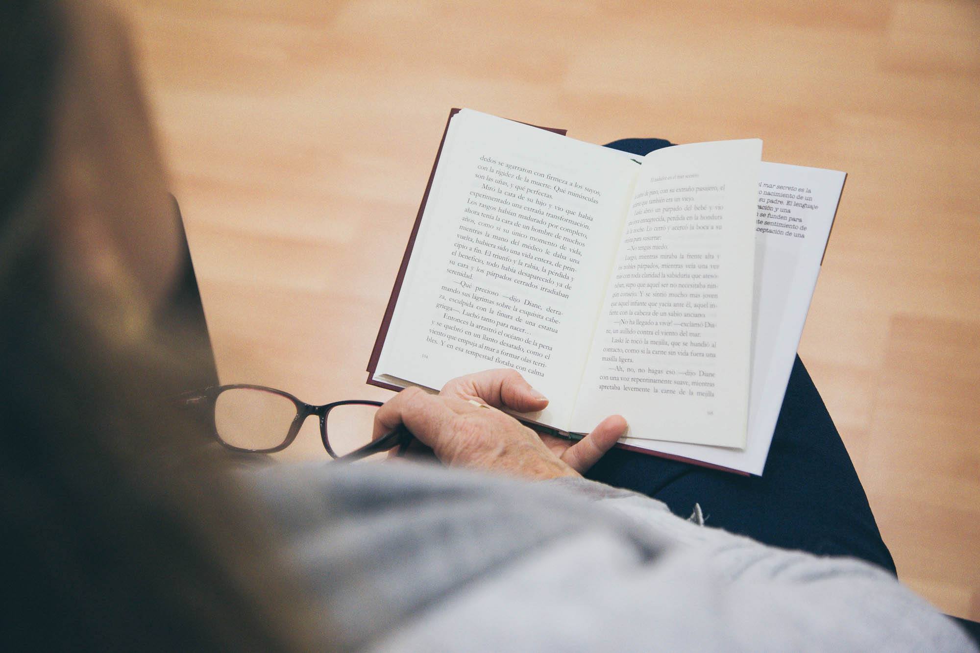 En la imagen aparece una persona leyendo un libro con una mano y en otra unas gafas de ver